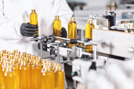 Automated process of production new cosmetic products on big modern factory. Scientist standing near conveyor line putting glass bottles with yellow thick substance on it. Production of new shampoo
