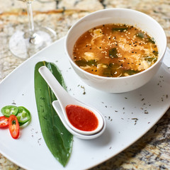 Korean cuisine. Spicy kimchi soup in white ceramic bowl served with red sauce in spoon, green leaf and paprika rings with glass of white wine on marble table.