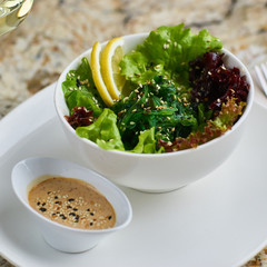 Seeweed salad in white ceramic bowl served with lemon slices, sauce, glass of white wine, fork and knife on marble table