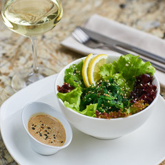 Seeweed salad in white ceramic bowl served with lemon slices, sauce, glass of white wine, fork and knife on marble table