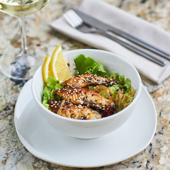 Asian cuisine. Warm salad with eel fish and rice served in white ceramic bowl with knife, fork and glass of white wine on marble background