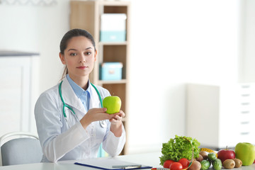 Young female nutritionist holding apple while sitting at table in her office