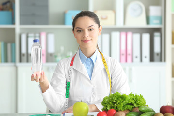 Young female nutritionist holding bottle of water while sitting at table in her office