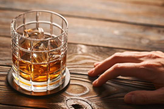 Glass of whiskey with ice cubes and man's hand on wooden table