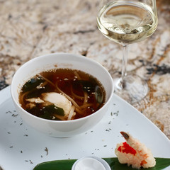 Japanese cuisine. Noodle soup udon in white ceramic bowl served with spoon, glass of white wine and king prawn on marble table