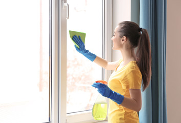 Young woman using detergent and rag while cleaning window in light room