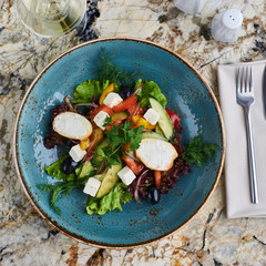 Greek salad with giant olives and provencal herbs served in blue ceramic plate with glass of white wine on marble table. Top view or flat lay.