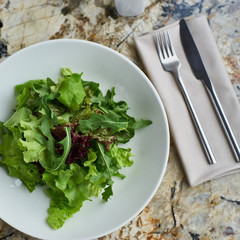 Lettuce salad served in white bowl with glass of white wine. Knife and fork lying on textile napkin on marble table. Top view or flat lay.