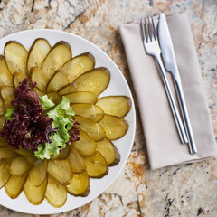 Slices ofmarinated cucumber on white plate. Knife and fork on textile napkin on marble table. Top view or flat lay.