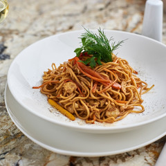 Flat egg noodles with vegetables served in big white ceramic round plate with glass of white wine on marble table