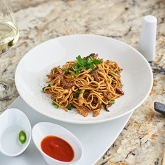 Flat egg noodles with duck meat, chicken, shiitake mushrooms served on white plate with white garlic and red spicy sauce