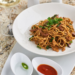 Flat egg noodles with duck meat, chicken, shiitake mushrooms served on white plate with white garlic and red spicy sauce