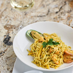 Flat egg noodles with seafood served in big white ceramic round plate with glass of white wine on marble table