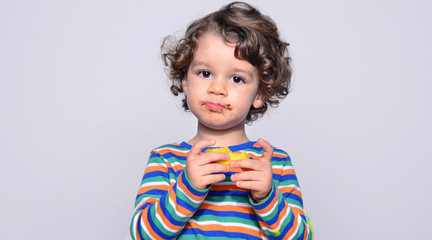 Kid getting messy while eating a chocolate cake. Beautiful curly hair boy eating sweets. Toddler in...