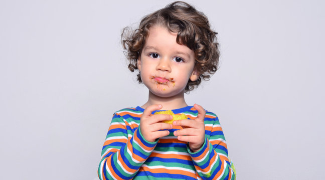 Kid getting messy while eating a chocolate cake. Beautiful curly hair boy eating sweets. Toddler in high chair being hungry stuffing his mouth with cake 
