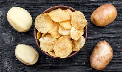 Bowl with potato chips and raw potatoes on the black wooden background.