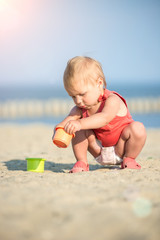 Baby playing on the sandy beach near the sea. Cute little girl in red dress with sand on tropical beach. Ocean coast.