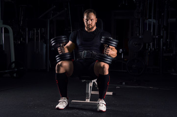 Impressive athlete sits on a bench with two dumbbells on his legs
