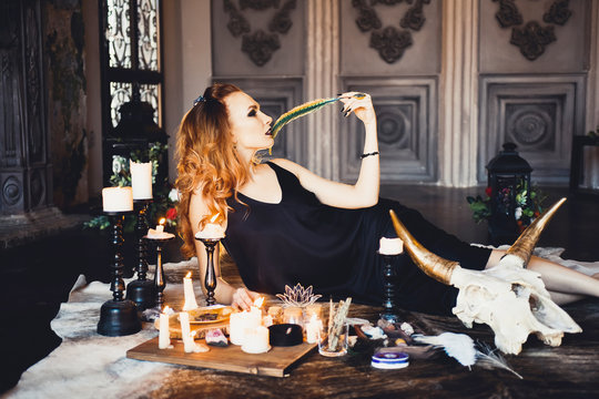 close-up portrait of young beautiful red-haired girl in the image of a Gothic witch on Halloween on a background of wax candles, fire, magic crystals and stones in the black dress conjures