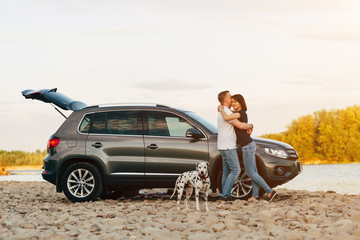 Family of two person and dog embrace near car on sunset river