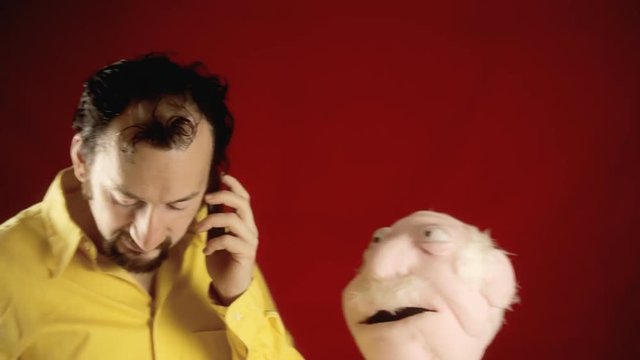 A funny ugly man talking on the phone, being disturbed by the puppet of an old man trying to stop the conversation.
