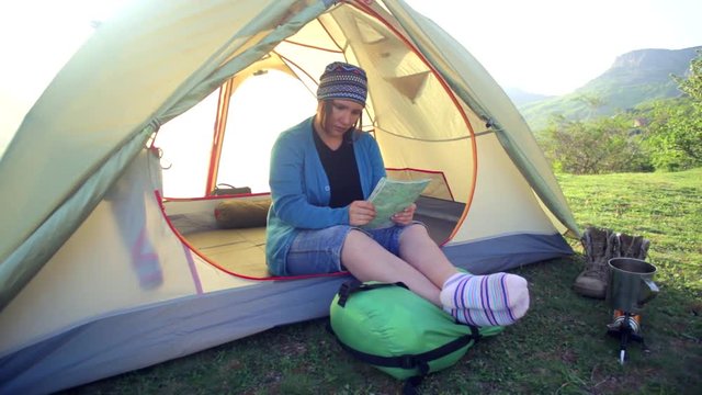 Woman in colorful socks and kerchief sits in a light yellow tent with sunlight shining through it and and turns over the map with travel boots and gas-burner standing near on green grass. 