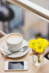 Cup of cappuccino, phone and bouquet of yellow flowers, wooden table