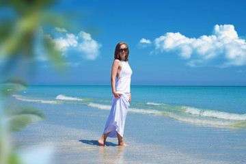 Beautiful girl in a long white dress walking on the beach with a clear water off the Caribbean bay.