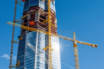 Scale active skyscraper building, glare from the sun on the windows, cranes against the blue sky.