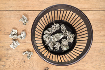 Trash can with crumpled dollars on a wooden surface