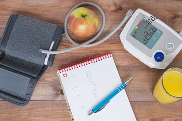 Healthy lifestyle concept, blood pressure monitor, notebook with measurement results and an apple on the brown wooden background.