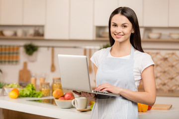Young woman standing near desk and laptop in the kitchen, smiling, looking at camera