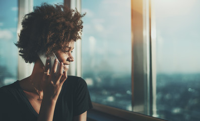 Portrait of charming Brazilian lady with perfect white smile sitting next to big windows of cruise ship and talking to her friend via smartphone, with stunning teal ocean in blurred background