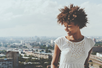 Cute laughing ebony girl looking down from balcony, smiling mixed teenage female with summer cityscape in background, young black lady in white dress with copy space zone for advertising or logo