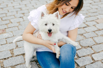 Beautiful young woman playing with her little west highland white terrier in a park outdoors. Lifestyle portrait.