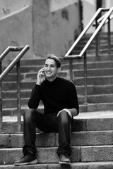 Young man talking on mobile while sitting on the stairs outdoors. Black-and-white photo.