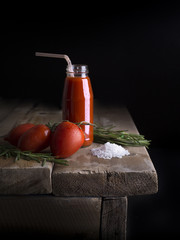 Tomato red juice in a glass bottle with fresh tomatoes, a sprig of rosemary and sea salt on a wooden old table on a black background. Side view. Сopy place. Mock up. Vegetable.