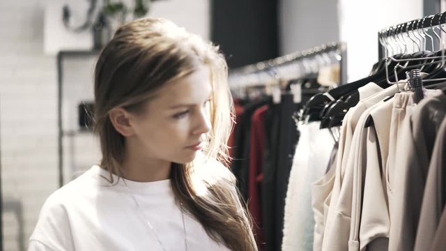 Beautiful young woman wearing a white T shirt is passing by racks of clothes a store trying to find what she wants and stopping near hoodies rack. Handheld real time close up shot