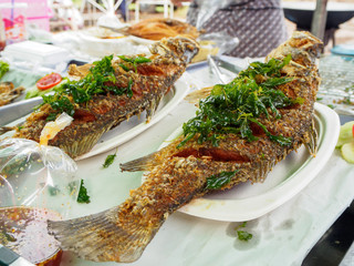 Close-up detail of multiple deep-fried sea bass fishes topped with herbs and spicy sauce at a local market. Chanthaburi, Thailand. Food and travel concept.