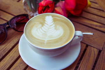 Cup of hot latte art coffee, glasses and tulips on wooden table