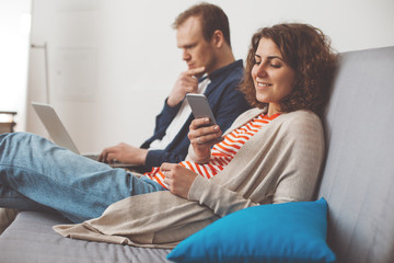 Couple spending time together at home. Man using computer and woman typing message with mobile phone. Sitting together on the sofa. Family life