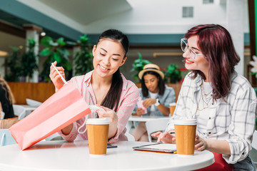 Smiling young women sitting at table and looking in shopping bag, young girls shopping concept