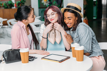 Smiling young women sitting at table with paper cups and holding new stylish shoes, young girls shopping concept