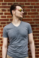 Pretty handsome young man hipster with sunglasses smiling laughing behind red brick wall. Looking aside.