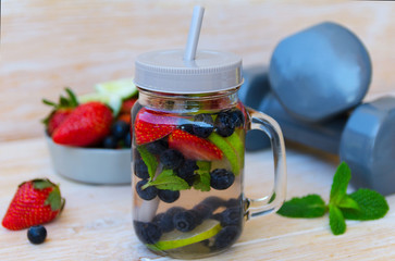 Fruit infused water with lime, lemons, strawberries, blueberries and mint in mason jar, grey dumbbells  and bowl with berries on wooden background. Healthy lifestyle, detox diet, loss weight concept.