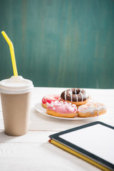 Unhealthy breakfast with coffee to go, plate of frosted donuts and digital tablet with white screen on wooden table. donuts and coffee background