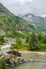 Fototapeta na wymiar Summer cloudy landscape with Russian Caucasus snowy and woody mountains and small river at the foot of it..
