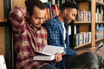 Young concentrated two african men reading books using laptop