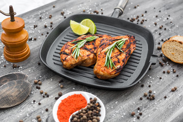 On a gray wooden surface lie spices, hand-made wooden pepper mill, grill pan, ready-made chicken breasts, rosemary, black pepper, large salt, red paprika, fresh bread, lime