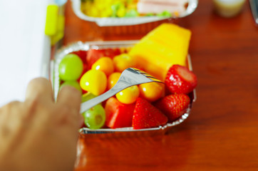 Healthy man using a fork to eat in the office a delicious fresh fruit salad on aluminium box on wooden table above view